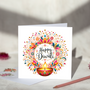 Happy Diwali greeting card with black script lettering surrounded by colourful floral patterns, gold decorative flourishes and a glowing diya, displayed with a white envelope